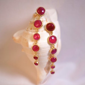 May include: A pair of long, dangling earrings with multiple pink gemstones and gold accents. The earrings are displayed on a seashell.