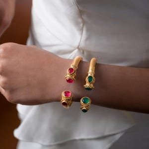 May include: A gold-coloured jewellery set, including a cuff bracelet and a ring, each embellished with a vivid red or green gemstone. The cuff bracelets have a rope-like design on the ends. The ring and bracelets are displayed on an arm.