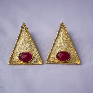 May include: A pair of gold-coloured triangle earrings with a textured surface. Each earring features a red oval-shaped stone in the centre. The earrings are displayed on a white surface.