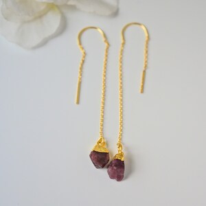 Könnte beinhalten: Goldfarbene Ohrhänger mit rohen, dunkelrosa Edelsteinen. Die Ohrringe haben ein schlichtes Design mit einer Kette und einem kleinen Stab oben. Die Edelsteine sind unregelmäßig geformt und haben eine goldfarbene Kappe.