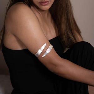 Può includere: Un braccialetto per braccio in argento con un design semplice e minimalista. Il braccialetto è composto da due sottili pezzi piatti di metallo che sono uniti alle estremità. Il braccialetto si indossa sulla parte superiore del braccio, appena sotto la spalla.