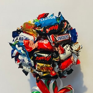 Puede incluir: Un arreglo de árbol de caramelos en un cubo rojo. El árbol está hecho de varias barras de chocolate envueltas, con un lazo colorido de cintas rojas, blancas y verdes. Una idea de regalo festiva y dulce.