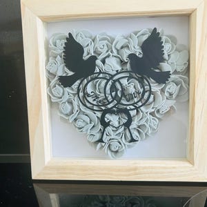 May include: A framed artwork with a heart of grey roses. Two black doves and interlocking rings are centered above the roses. The word "Mr" is visible on the rings. The frame is made of light-coloured wood.