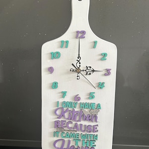 Puede incluir: Reloj de cocina de madera blanca con forma de tabla de cortar. El reloj tiene números turquesa y morados, con el texto "I only have a kitchen because it came with the house." Las manecillas del reloj son negras.
