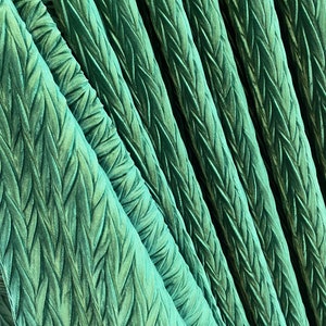 May include: A vibrant emerald green fabric featuring a repeating, raised herringbone or braided texture. The material is gathered into deep, vertical folds, creating a series of diagonal ridges that catch the light and emphasize the pattern.