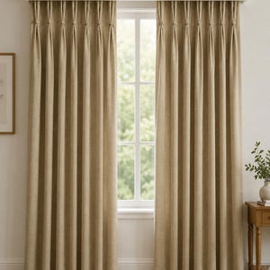 May include: A pair of floor-length, beige curtains featuring a pinch pleat header design. The fabric has a textured, woven appearance and hangs in uniform vertical folds from a white curtain rod mounted above a window frame.