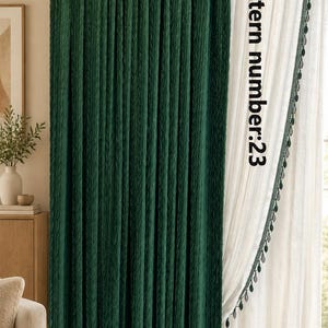 May include: A dark green curtain with a textured, pleated surface hangs from a gold rod next to a sheer white curtain featuring a decorative green pom-pom trim. The text colour pattern number:23 is printed vertically on the white curtain.