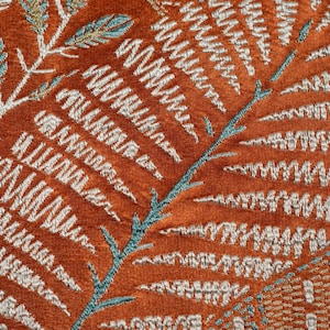 May include: Close-up of a textured fabric featuring an embroidered fern pattern. The background is a warm orange-brown, with the fern design in white and teal. The detailed embroidery creates a tactile and visually rich surface.