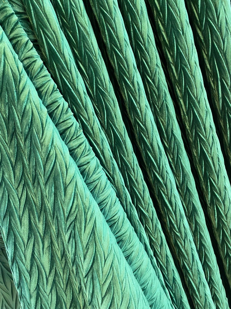 May include: A vibrant emerald green fabric featuring a repeating, raised herringbone or braided texture. The material is gathered into deep, vertical folds, creating a series of diagonal ridges that catch the light and emphasize the pattern.