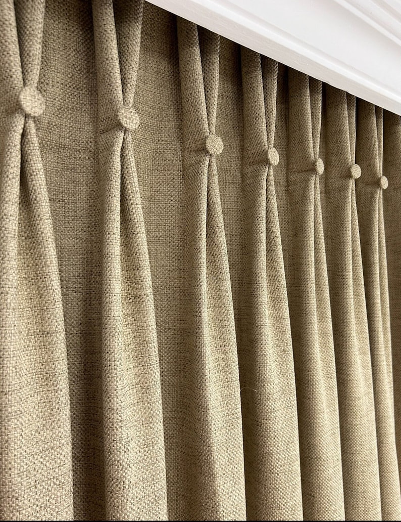May include: A beige curtain with a textured fabric finish featuring a series of vertical pleats. Each pleat is secured at the top with a small, matching fabric-covered button. The curtain is suspended from a white horizontal track system.