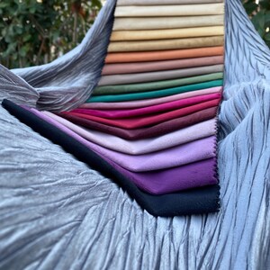 May include: A fabric swatch sample book featuring a vertical stack of rectangular velvet-like material strips in a gradient of colours including beige, yellow, orange, green, pink, red, purple, and black, resting on a crinkled silver-grey fabric.