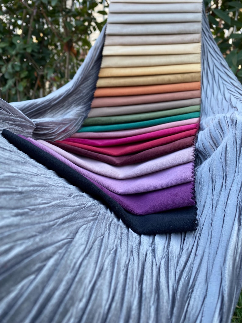 May include: A fabric swatch sample book featuring a vertical stack of rectangular velvet-like material strips in a gradient of colours including beige, yellow, orange, green, pink, red, purple, and black, resting on a crinkled silver-grey fabric.