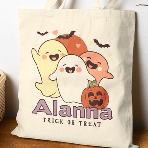 Könnte beinhalten: Beige Canvas-Tragetasche mit einem Halloween-Design. Das Design zeigt vier lächelnde Geister, einen Kürbis und Fledermäuse. Der Name "Alanna" und der Satz "Trick or Treat" sind darunter aufgedruckt.