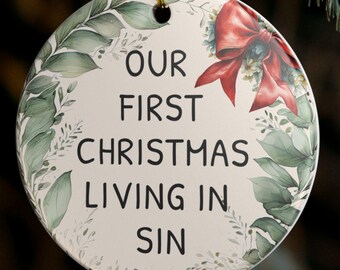May include: Round Christmas ornament with the text "OUR FIRST CHRISTMAS LIVING IN SIN". The ornament has a red bow and green leaf design. It is hanging from a gold string.