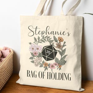 以下が含まれることがあります： 「Stephanie's BAG OF HOLDING」の文字と花のリースデザインが施された、クリーム色のトートバッグ。 リースは黒い20面ダイスを囲んでいます。 バッグは長いハンドル付きで、キャンバスのような素材で作られています。