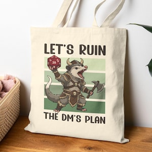 Könnte beinhalten: Beige Canvas-Tragetasche mit einem Cartoon-Opossum-Charakter, der einen roten zwanzigseitigen Würfel und eine Axt hält. Der Text "LET'S RUIN THE DM'S PLAN" ist oberhalb und unterhalb der Illustration aufgedruckt. Die Tasche hat lange Griffe.
