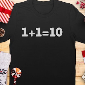 Könnte beinhalten: Schwarzes T-Shirt mit dem weißen Text "1+1=10". Das Shirt wird auf einer Holzoberfläche mit Weihnachtsdekorationen präsentiert, darunter ein verpacktes Geschenk, Ornamente und ein Schneeflocke.