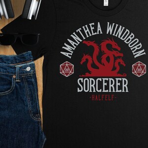 Könnte beinhalten: Schwarzes T-Shirt mit einem roten Drachen und dem Text "Amathea Windborn Sorcerer -Halfel-". Der Drache ist von zwei weißen 20-seitigen Würfeln umgeben.