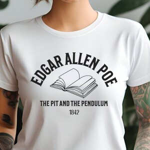 Könnte beinhalten: Weißes T-Shirt mit einem schwarzen Grafikdesign, das den Text "Edgar Allen Poe" in einem Bogen über einer Illustration eines offenen Buches zeigt. Der Text "The Pit and the Pendulum 1842" befindet sich unter dem Buch.