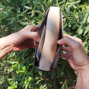 Puede incluir: Una cartera de cuero marr&oacute;n con m&uacute;ltiples ranuras para tarjetas y un portafolios. La cartera est&aacute; abierta y el interior est&aacute; forrado con una tela beige.