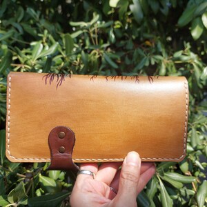 Puede incluir: Una cartera de cuero marr&oacute;n con cierre de solapa y un dise&ntilde;o decorativo. La cartera tiene costuras blancas alrededor de los bordes y una correa de cuero marr&oacute;n con dos remaches met&aacute;licos.