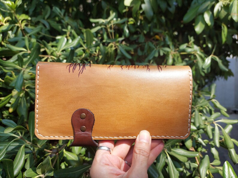 Puede incluir: Una cartera de cuero marr&oacute;n con cierre de solapa y un dise&ntilde;o decorativo. La cartera tiene costuras blancas alrededor de los bordes y una correa de cuero marr&oacute;n con dos remaches met&aacute;licos.