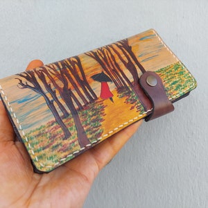 Puede incluir: Una cartera de cuero marr&oacute;n con una escena pintada de una mujer caminando por un bosque con un paraguas. La cartera tiene una correa de cuero marr&oacute;n con un bot&oacute;n de metal.