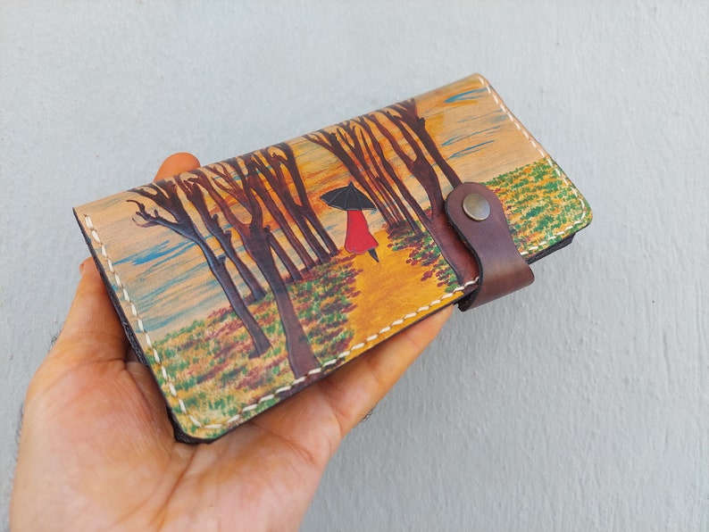 Puede incluir: Una cartera de cuero marr&oacute;n con una escena pintada de una mujer caminando por un bosque con un paraguas. La cartera tiene una correa de cuero marr&oacute;n con un bot&oacute;n de metal.