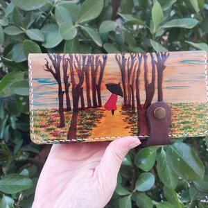 Puede incluir: Una cartera de cuero marr&oacute;n con una escena pintada de una persona caminando por un bosque con un paraguas. La escena est&aacute; pintada en tonos marrones, verdes, amarillos y azules.