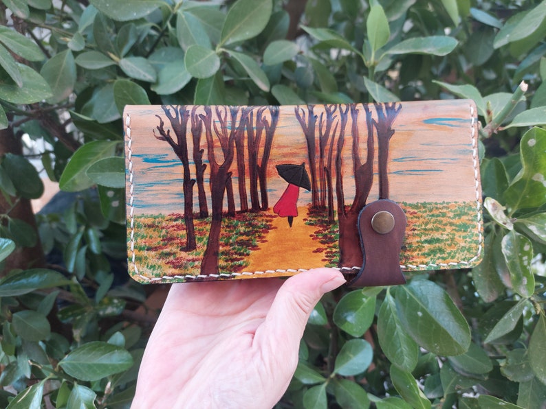 Puede incluir: Una cartera de cuero marr&oacute;n con una escena pintada de una persona caminando por un bosque con un paraguas. La escena est&aacute; pintada en tonos marrones, verdes, amarillos y azules.