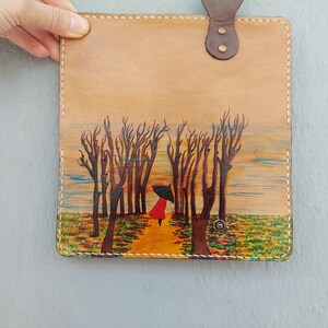 Puede incluir: Una cubierta de cuaderno de cuero marr&oacute;n con una escena pintada de una persona de rojo caminando por un bosque con un paraguas. El fondo es una puesta de sol con un cielo azul y nubes amarillas.
