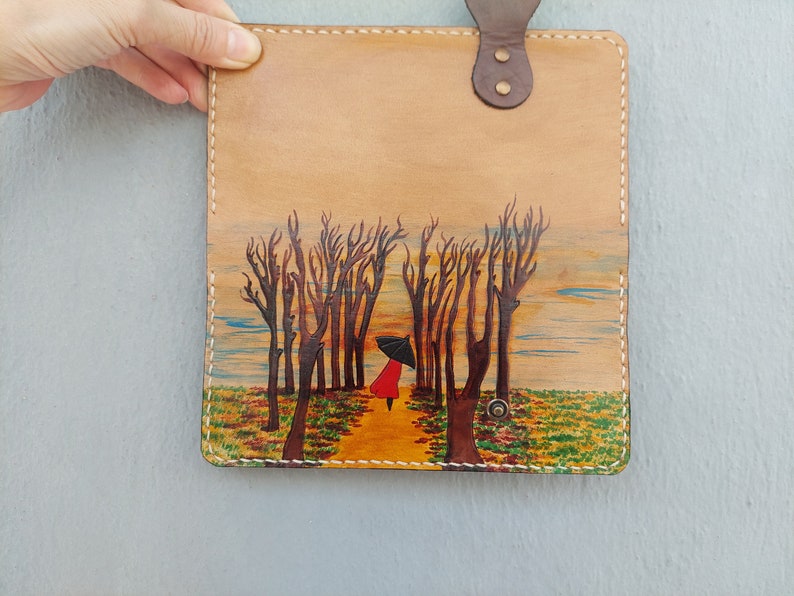 Puede incluir: Una cubierta de cuaderno de cuero marr&oacute;n con una escena pintada de una persona de rojo caminando por un bosque con un paraguas. El fondo es una puesta de sol con un cielo azul y nubes amarillas.