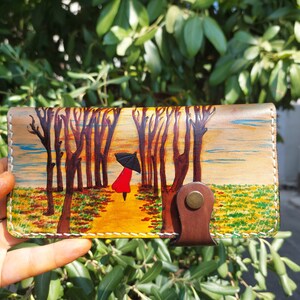 Puede incluir: Una cartera de cuero con una escena pintada de una persona caminando bajo un paraguas en un bosque. La cartera tiene una solapa de cuero marr&oacute;n con un bot&oacute;n de lat&oacute;n.