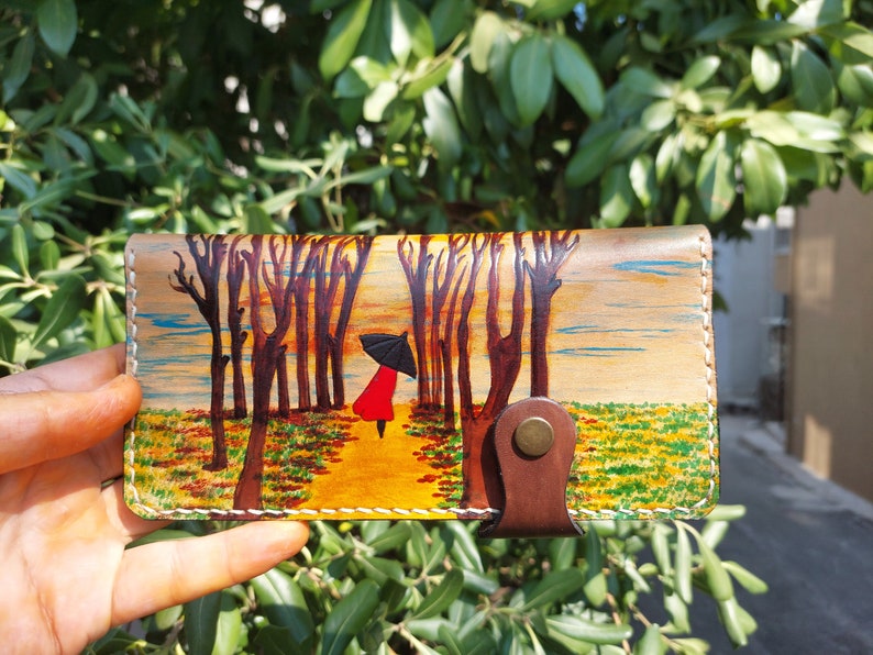 Puede incluir: Una cartera de cuero con una escena pintada de una persona caminando bajo un paraguas en un bosque. La cartera tiene una solapa de cuero marr&oacute;n con un bot&oacute;n de lat&oacute;n.