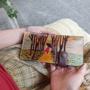 Puede incluir: Una cartera de cuero marr&oacute;n con una escena pintada de una mujer caminando por un bosque con &aacute;rboles a cada lado. La mujer lleva un abrigo rojo y lleva una sombrilla. La cartera tiene una solapa de cuero marr&oacute;n con cierre de presi&oacute;n.