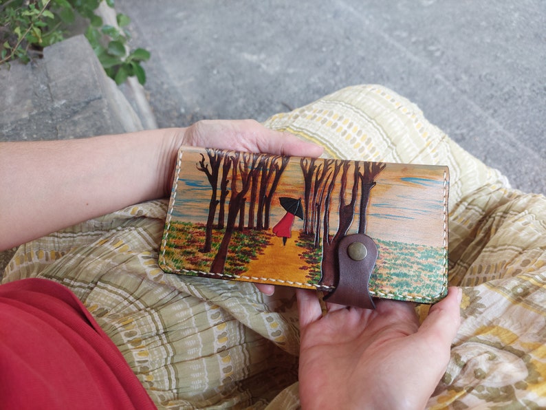 Puede incluir: Una cartera de cuero marr&oacute;n con una escena pintada de una mujer caminando por un bosque con &aacute;rboles a cada lado. La mujer lleva un abrigo rojo y lleva una sombrilla. La cartera tiene una solapa de cuero marr&oacute;n con cierre de presi&oacute;n.