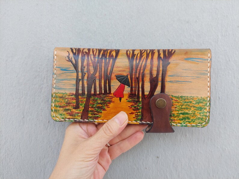 Puede incluir: Una cartera de cuero marr&oacute;n con una escena pintada de una persona caminando por un bosque con un paraguas. La escena est&aacute; pintada en tonos marrones, amarillos, verdes y rojos.