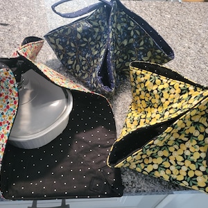 May include: Three fabric casserole carriers with handles, featuring different patterns: one with colorful flowers, one with blue berries and leaves, and one with yellow lemons. Each carrier has a black interior lining with white polka dots.