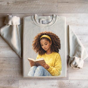 Puede incluir: Una sudadera beige con una ilustraci&oacute;n de una persona con cabello casta&ntilde;o rizado, que lleva una diadema y un su&eacute;ter amarillos, leyendo un libro. La persona lleva vaqueros azules. La sudadera est&aacute; sobre una superficie de madera.