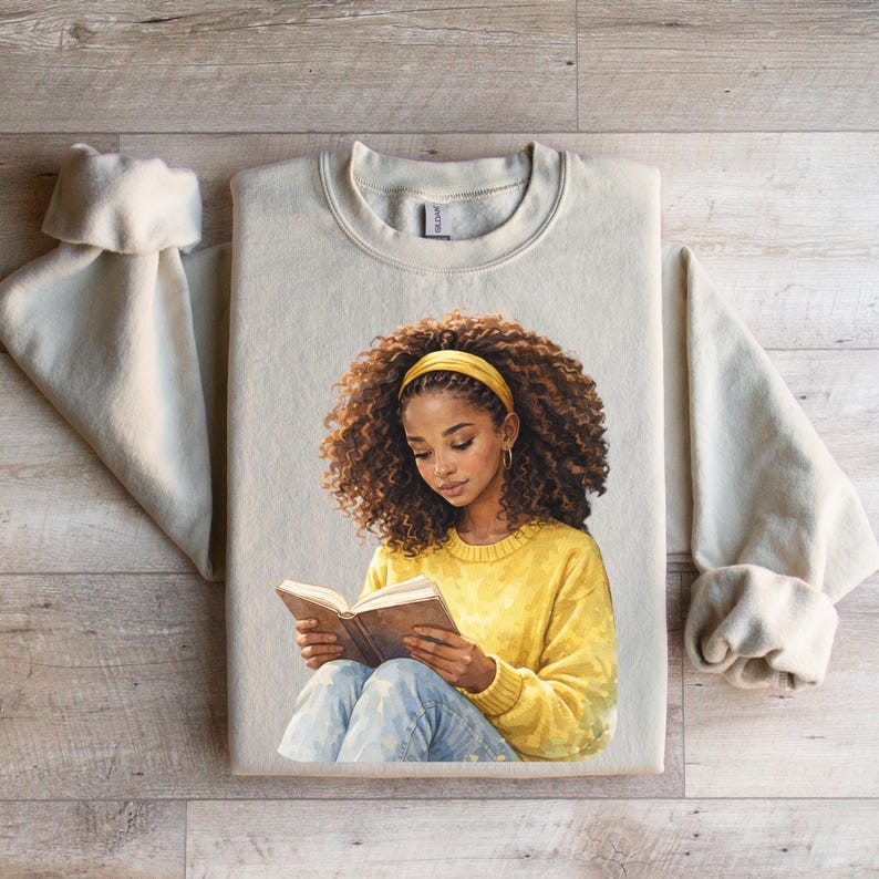Puede incluir: Una sudadera beige con una ilustraci&oacute;n de una persona con cabello casta&ntilde;o rizado, que lleva una diadema y un su&eacute;ter amarillos, leyendo un libro. La persona lleva vaqueros azules. La sudadera est&aacute; sobre una superficie de madera.