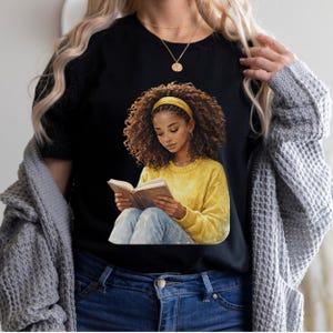 Puede incluir: Camiseta negra con una ilustraci&oacute;n en acuarela de una persona leyendo un libro. La ilustraci&oacute;n muestra a una persona con cabello casta&ntilde;o rizado, que lleva un su&eacute;ter amarillo, jeans azules y una diadema amarilla. Tambi&eacute;n se ve un collar dorado.