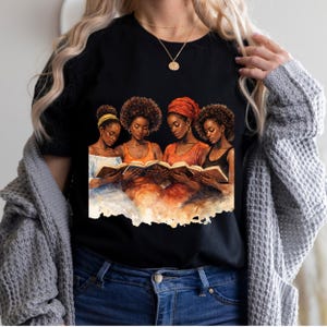 Puede incluir: Camiseta negra con una ilustraci&oacute;n en acuarela de cuatro mujeres leyendo libros. Las mujeres, de piel oscura, visten ropa naranja, amarilla y negra, con tocados. Se ve un collar dorado.
