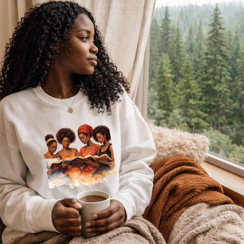 Puede incluir: Una sudadera blanca con una ilustraci&oacute;n en acuarela de cuatro mujeres negras leyendo un libro. Las mujeres tienen diferentes peinados y ropa. La sudadera la lleva una persona que sostiene una taza de caf&eacute;, sentada cerca de una ventana con vista al bosque.