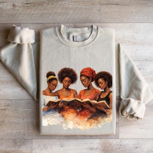 Puede incluir: Sudadera color crema con cuello redondo y una ilustraci&oacute;n en acuarela de cuatro mujeres leyendo libros. Las mujeres tienen piel oscura y varios peinados, una de ellas con un pa&ntilde;uelo en la cabeza. La obra de arte est&aacute; en tonos c&aacute;lidos de naranja, marr&oacute;n y azul.