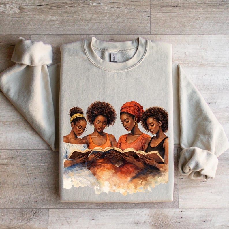 Puede incluir: Sudadera color crema con cuello redondo y una ilustraci&oacute;n en acuarela de cuatro mujeres leyendo libros. Las mujeres tienen piel oscura y varios peinados, una de ellas con un pa&ntilde;uelo en la cabeza. La obra de arte est&aacute; en tonos c&aacute;lidos de naranja, marr&oacute;n y azul.