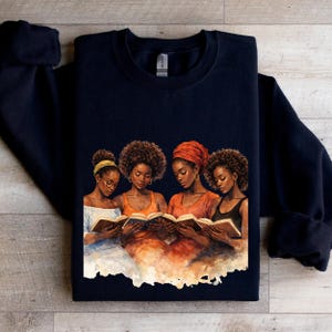 Puede incluir: Sudadera azul marino con una ilustraci&oacute;n en acuarela de cuatro mujeres leyendo libros. Las mujeres tienen piel oscura y visten tocados y atuendos. La obra de arte est&aacute; en tonos c&aacute;lidos de naranja, marr&oacute;n y amarillo.