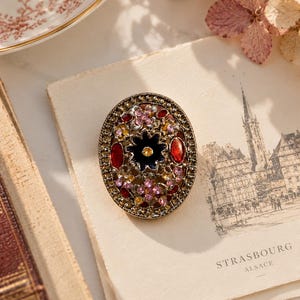 Puede incluir: Un broche de forma ovalada con un marco de filigrana de metal oscuro engastado con cristales multicolores y detalles en esmalte. El centro muestra un motivo de flor negra rodeado de piedras rojas, rosas y amarillas, con un borde texturizado.