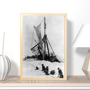 May include: A framed black and white photograph depicting a ship wrecked in a snowy landscape. The ship's mast is broken, and several figures are seen in the snow. The photograph is in a gold frame.