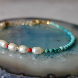 May include: A delicate bracelet with white pearls, red coral beads, and turquoise beads. The bracelet is on a blue and white agate slice.