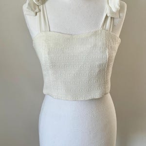 Peut inclure: Haut court crème en tissu texturé. Il présente un col carré et des bretelles ornées de nœuds. Le vêtement est présenté sur un mannequin blanc, mettant en valeur son design. Le style est élégant et adapté à diverses occasions. Le tissu est probablement tissé.
